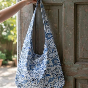 Könnte beinhalten: Eine blau-weiß gemusterte Umhängetasche. Die Tasche ist aus Stoff und hat einen langen Riemen. Das Blumenmuster zeigt blaue Blumen und Blätter auf weißem Hintergrund. Die Tasche wird von einer Hand gehalten.