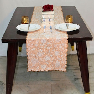 Puede incluir: Una mesa de comedor puesta para una comida. Un camino de mesa floral color melocotón con borde festoneado recorre el centro de la mesa de madera oscura. Platos blancos, cubiertos y candelabros dorados están a ambos lados. Una caja espejada y flores rojas están en el centro.
