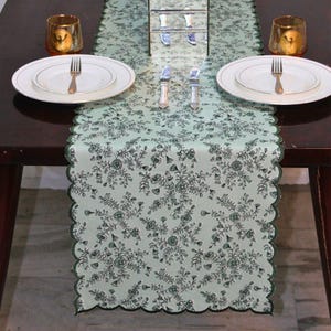 Op de afbeelding: Lichtgroene bloemen tafelloper met geschulpte rand, op een donkerhouten tafel. De loper heeft een herhalend patroon van bloemen en bladeren in een donkerdere groene kleur. Twee witte borden, vorken en messen staan op de tafel.