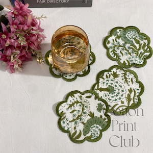 Peut inclure: Un verre d'eau est posé sur un dessous de verre à motifs floraux et bordure festonnée. Trois autres dessous de verre sont visibles, avec un fond blanc, des motifs floraux verts et une bordure verte. Collection Cotton Print Club.