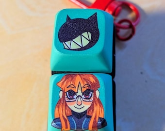 May include: A teal keychain featuring two stacked square keycaps. The top keycap has a black cat face design, and the bottom keycap has a cartoon image of a person with orange hair and glasses. A red clasp is attached.