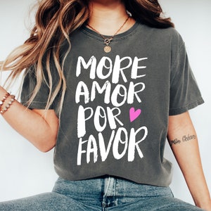May include: A grey t-shirt with the text "More Amor Por Favor" in white with a pink heart. The shirt is being worn by a person with long brown hair.