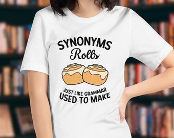 Puede incluir: Camiseta blanca con el texto "Synonyms Rolls" sobre una ilustración de dos rollos de canela. Debajo de los rollos, el texto dice "Just like grammar used to make". La camiseta la lleva una persona frente a una estantería.