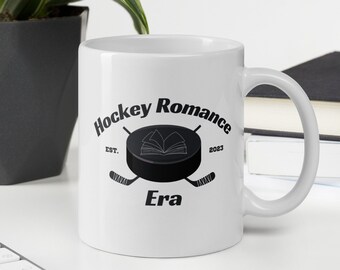 May include: White ceramic mug with the words "Hockey Romance Era" in black, featuring a hockey puck and crossed hockey sticks design. The mug also includes the text "Est. 2023". The mug is on a white surface.