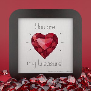 May include: A framed cross-stitch design with a red heart-shaped gem and the text "You are my treasure!" The background is white and the frame is black.