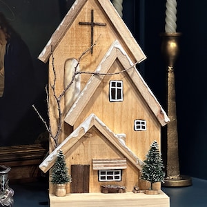 May include: A wooden church-shaped decorative piece with a cross on the roof. The structure has multiple levels, with a small door, windows, and miniature evergreen trees. The roof edges are trimmed with white accents, and a small branch is attached to the side.