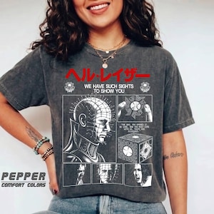 May include: A charcoal grey short-sleeve t-shirt featuring a white graphic print of Pinhead from Hellraiser. The design includes red Japanese text, the phrase WE HAVE SUCH SIGHTS TO SHOW YOU, and illustrations of the puzzle box and character.