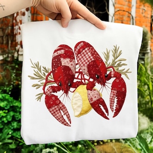 May include: White sweatshirt with a graphic of two red crawfish, featuring patterned details, a yellow lemon slice, and sprigs of greenery. The crawfish have a patchwork design. The sweatshirt is held by a hand.