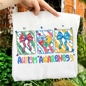 Puede incluir: Sudadera blanca con un gráfico colorido que promueve la concienciación sobre el autismo. El diseño presenta tres imágenes enmarcadas con un lazo rojo, un rompecabezas en forma de corazón y un lazo azul. Las palabras "AUTISM AWARENESS" están escritas en colores.