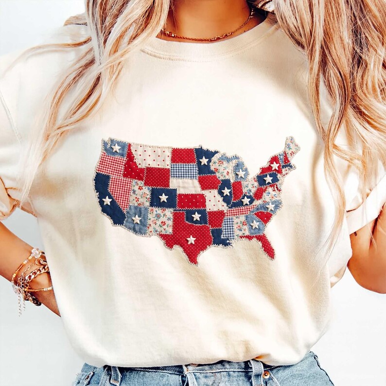 May include: Cream-colored t-shirt featuring a patchwork design of the United States map. The map is composed of red, white, and blue fabric squares with star accents. The shirt is a casual, patriotic style.
