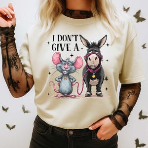 Puede incluir: Camiseta color crema con un rat&oacute;n y un burro de dibujos animados con el texto "I DON'T GIVE A." El rat&oacute;n es gris con orejas rosas, y el burro es negro. Ambos llevan accesorios rosas. La camiseta tiene cuello redondo y mangas cortas.