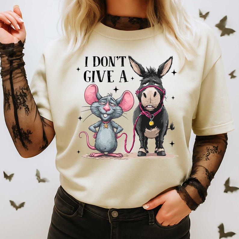 Puede incluir: Camiseta color crema con un rat&oacute;n y un burro de dibujos animados con el texto "I DON'T GIVE A." El rat&oacute;n es gris con orejas rosas, y el burro es negro. Ambos llevan accesorios rosas. La camiseta tiene cuello redondo y mangas cortas.