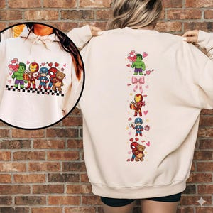 May include: A cream-colored sweatshirt featuring cartoon versions of Marvel superheroes. The front displays Hulk, Iron Man, Captain America, and Spider-Man with hearts and balloons. The back has a vertical design with the same characters and bows.
