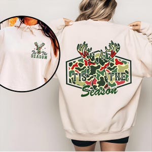 May include: Cream-colored sweatshirt with a festive design. The back features a camouflage-style deer head with red and green accents, and the text "Tis The Season." A smaller version of the design is on the front of the hoodie.