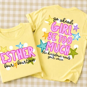 以下が含まれることがあります： カラフルなグラフィックが施された淡い黄色のTシャツ2枚。1枚にはピンクと黒で「ESTHER four:fourteen」という文字と、黄色、青、緑の星が描かれています。もう1枚には「go ahead, GIRL BE TOO MUCH the kingdom needs your voice.」と書かれています。