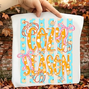 Könnte beinhalten: Ein weißes T-Shirt mit einem Grafikdruck aus vertikalen hellblauen Streifen, orangefarbenem, gepunktetem Text mit der Aufschrift COZY SEASON, orangefarbenen Kürbisillustrationen und zwei rosa-weiß karierten Schleifen an den Ecken.