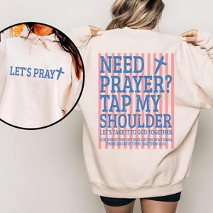 May include: Cream-colored sweatshirt with blue text that says "LET'S PRAY" and a cross. The back of the sweatshirt has the text "NEED PRAYER? TAP MY SHOULDER" in blue, with a cross graphic and pink stripes. The text "LET'S TAKE IT TO GOD TOGETHER" is also visible.