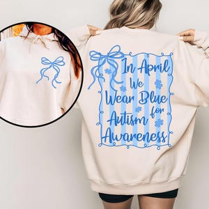 May include: A cream-colored sweatshirt with a blue bow design on the front and back. The back of the sweatshirt features the text "In April We Wear Blue Autism for Awareness" in blue, with blue puzzle pieces and vertical stripes.