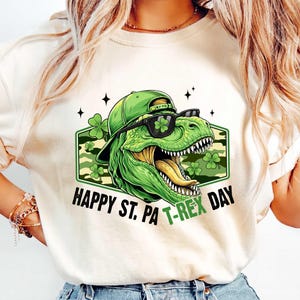 May include: Cream-coloured t-shirt with a green dinosaur wearing a hat and sunglasses, and the text "HAPPY ST. PA T-REX DAY". The dinosaur is inside a green and camouflage-patterned hexagon with shamrocks.