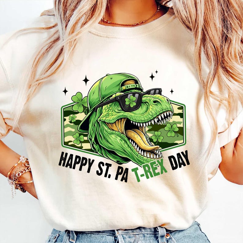 May include: Cream-coloured t-shirt with a green dinosaur wearing a hat and sunglasses, and the text "HAPPY ST. PA T-REX DAY". The dinosaur is inside a green and camouflage-patterned hexagon with shamrocks.