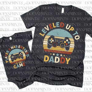 May include: A set of two gray shirts with a retro video game theme. The larger shirt says "Leveled Up To Daddy" with a vintage-style video game controller and a sunset background. The smaller shirt says "Player 2 Has Entered The Game" with a similar video game controller and sunset background.