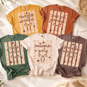 May include: Five short-sleeved t-shirts in mustard yellow, burnt orange, sage green, cream, and dark brown. Each shirt features a central rectangular graphic with vertical stripes, floral patterns, stars, a bow icon, and seasonal text.