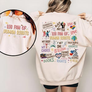 May include: A cream-coloured hoodie featuring the text "BIG FAN OF HUMAN RIGHTS" on the front. The back displays a graphic with phrases and symbols, including "Black Lives Matter" and a rainbow heart, promoting social justice and equality.