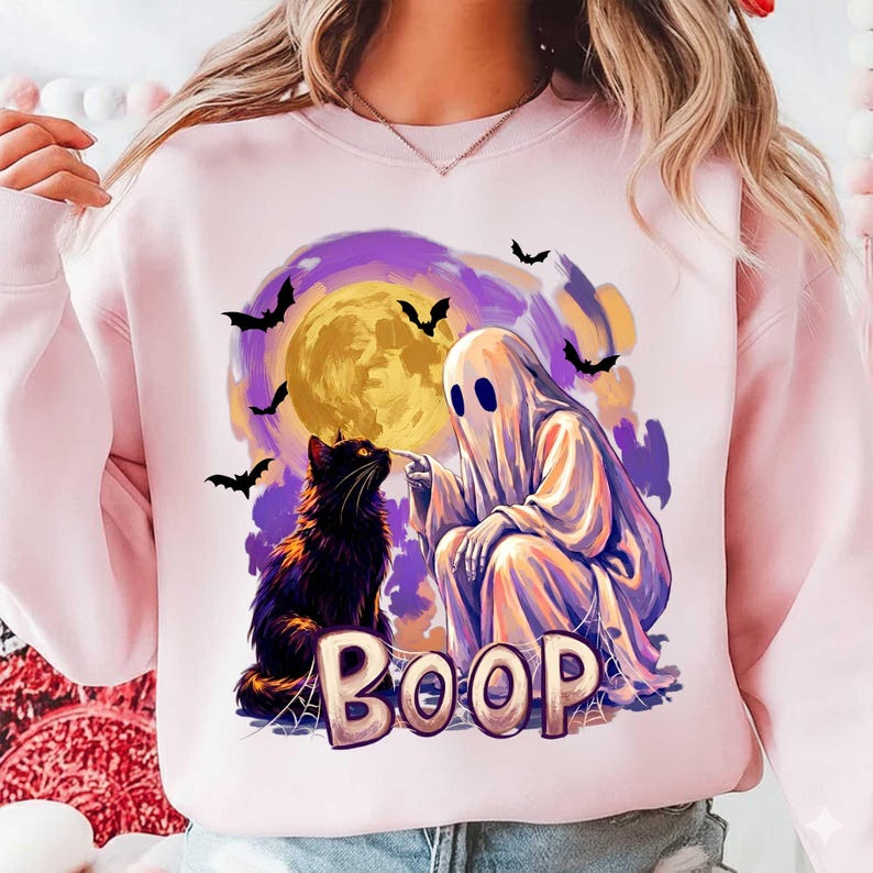 May include: A light pink sweatshirt featuring a graphic print of a white ghost touching the nose of a black cat. The design includes a large yellow moon, purple clouds, flying bats, and the word BOOP in large letters at the bottom.
