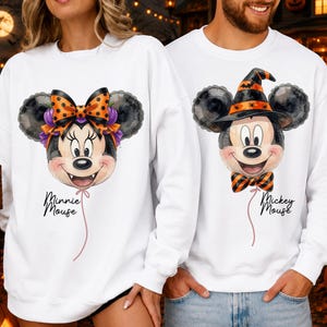 May include: Two white crewneck sweatshirts featuring watercolor-style balloon graphics. The left sweatshirt displays Minnie Mouse with a polka-dot bow and purple pumpkins, labeled Minnie Mouse. The right shows Mickey Mouse in a witch hat, labeled Mickey Mouse.