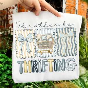 以下が含まれることがあります： 「I'd rather be thrifting」の文字が入った白いスウェットシャツ。デザインには、リボン、食器でいっぱいのショッピングカート、別のリボンの3つのフレーム入りイラストが含まれています。イラストは青、黄、白の色合いです。