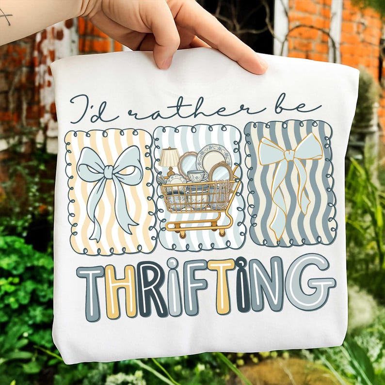 May include: White sweatshirt with the text "I'd rather be thrifting." The design includes three framed illustrations: a bow, a shopping cart filled with dishes, and another bow. The illustrations are in shades of blue, yellow, and white.