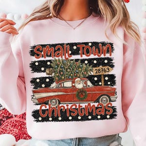 May include: A light pink crewneck sweatshirt featuring a graphic of a red vintage car carrying a decorated Christmas tree on its roof. Santa Claus is in the driver seat. The text reads "SMALL TOWN CHRISTMAS" with a signpost marked "28763".