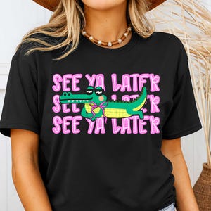 May include: Black t-shirt with the phrase "SEE YA LATER" in pink, repeated three times. A cartoon alligator wearing sunglasses and a pink bow tie is in the center. The alligator is green and yellow.