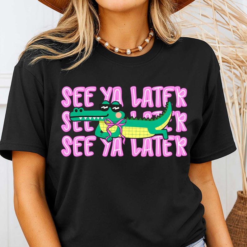 May include: Black t-shirt with the phrase "SEE YA LATER" in pink, repeated three times. A cartoon alligator wearing sunglasses and a pink bow tie is in the center. The alligator is green and yellow.