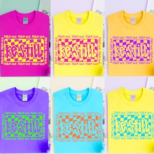 May include: A collection of six t-shirts in various colors: pink, yellow, purple, light blue, and two shades of yellow. Each shirt features the text "Be Still" in a checkered box, with "Psalm 46:10" printed around the box.