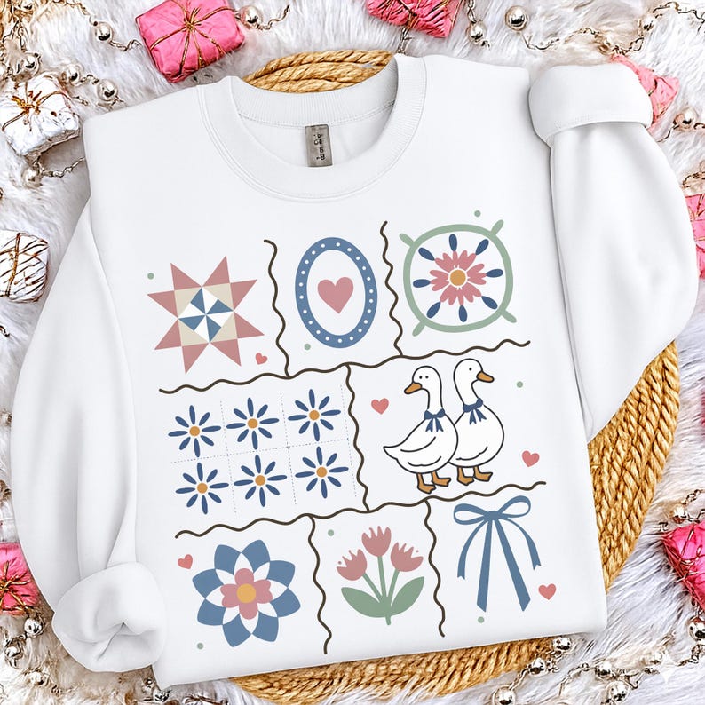 May include: White sweatshirt featuring a patchwork design with floral, heart, and duck illustrations. The design includes a star, a heart within a circle, and a bow. The sweatshirt is a cozy, casual garment.