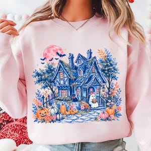Pu&ograve; includere: Una felpa girocollo rosa chiaro con un'illustrazione grafica di una casa stregata blu, una grande luna rosa, pipistrelli, zucche, un gatto nero e un piccolo fantasma con un fiocco rosa, circondati da fiori e fogliame colorato.