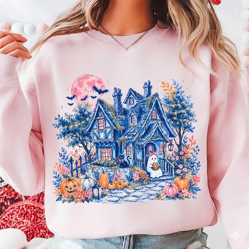Pu&ograve; includere: Una felpa girocollo rosa chiaro con un'illustrazione grafica di una casa stregata blu, una grande luna rosa, pipistrelli, zucche, un gatto nero e un piccolo fantasma con un fiocco rosa, circondati da fiori e fogliame colorato.