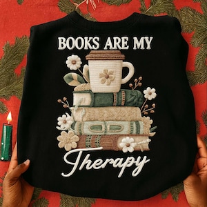 Może przedstawiać: Czarna bluza z haftowanym napisem "BOOKS ARE MY Therapy". Projekt przedstawia stos książek, filiżankę kawy i kwiatowe akcenty w kolorze białym, zielonym i brązowym.