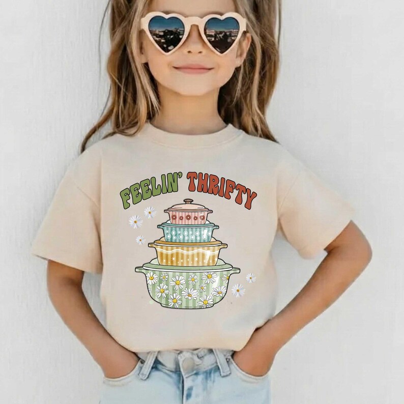 May include: Beige t-shirt with the text "FEELIN' THRIFTY" in green and red. The shirt features a stack of colorful, vintage-style casserole dishes with daisy accents. A child is wearing the shirt.