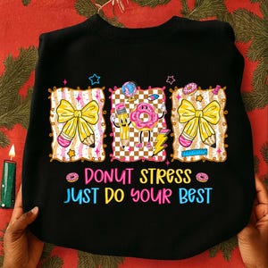 Puede incluir: Sudadera negra con un gráfico colorido que presenta un lazo amarillo, un lápiz y un personaje de donut rosa. El texto "DONUT STRESS JUST DO YOUR BEST" está impreso debajo en rosa y azul.