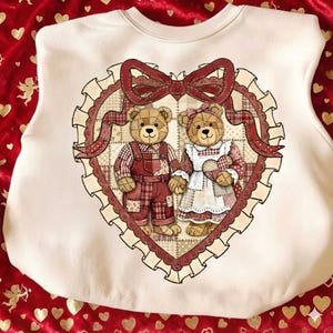 May include: Cream-colored sweatshirt featuring a heart-shaped design with two teddy bears. The bears are dressed in patchwork outfits, surrounded by a red bow and a scalloped border. The design is centered on the front of the sweatshirt.