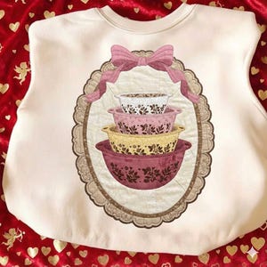 May include: Cream-colored sweatshirt featuring a decorative oval design with a stack of four patterned mixing bowls in shades of white, pink, yellow, and burgundy. A pink bow adorns the top of the oval. The design is framed by a scalloped border.