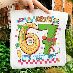 Puede incluir: Camiseta blanca con un diseño colorido. El diseño incluye la palabra "BRUH", los números "67" y el texto "IT'S TEST DAY". El diseño también presenta un lápiz, una manzana, una regla, libros y flores.