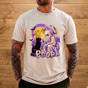 May include: A beige short-sleeve t-shirt featuring a graphic print of a white ghost touching the nose of a black cat. The design includes a large yellow full moon, flying bats, and the word BOOP in large purple letters centered below the figures.