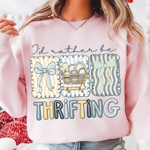 May include: Pink sweatshirt with the text "I'd rather be thrifting." The design includes illustrations of bows, a shopping cart filled with dishes, and striped patterns. The word "thrifting" is in a playful font.