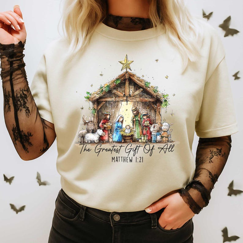 May include: Cream-colored t-shirt featuring a watercolor illustration of a nativity scene. The image includes a wooden stable, figures of Mary, Joseph, and baby Jesus, and the text "The Greatest Gift Of All" and "Matthew 1:21".