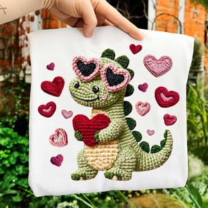May include: White tote bag featuring a crocheted dinosaur wearing heart-shaped sunglasses and holding a red heart. Surrounding the dinosaur are various crocheted hearts in shades of red, pink, and purple. The design is whimsical and handmade.