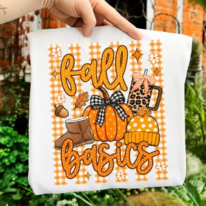 May include: A white t-shirt featuring an orange and white gingham background with the text fall basics in orange polka-dot script. The design includes an orange pumpkin with a black and white plaid bow, a brown boot, a leopard print mug, and a beanie.