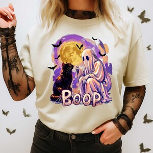 May include: A cream-colored t-shirt featuring a graphic of a white ghost touching the nose of a black cat. The design includes a large yellow moon, purple clouds, flying bats, and the word BOOP in large, light-colored, blocky letters.